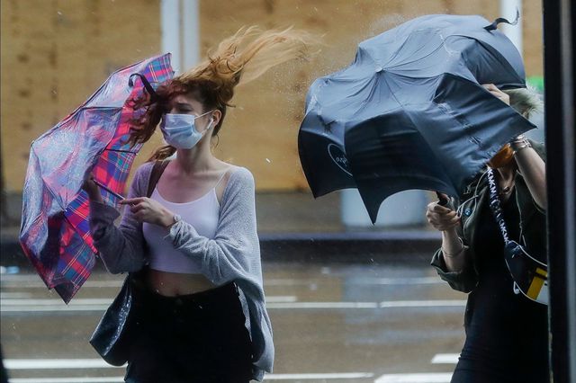 A woman in a mask struggles with an umbrella that is being blow inside out on July 10, 2020
